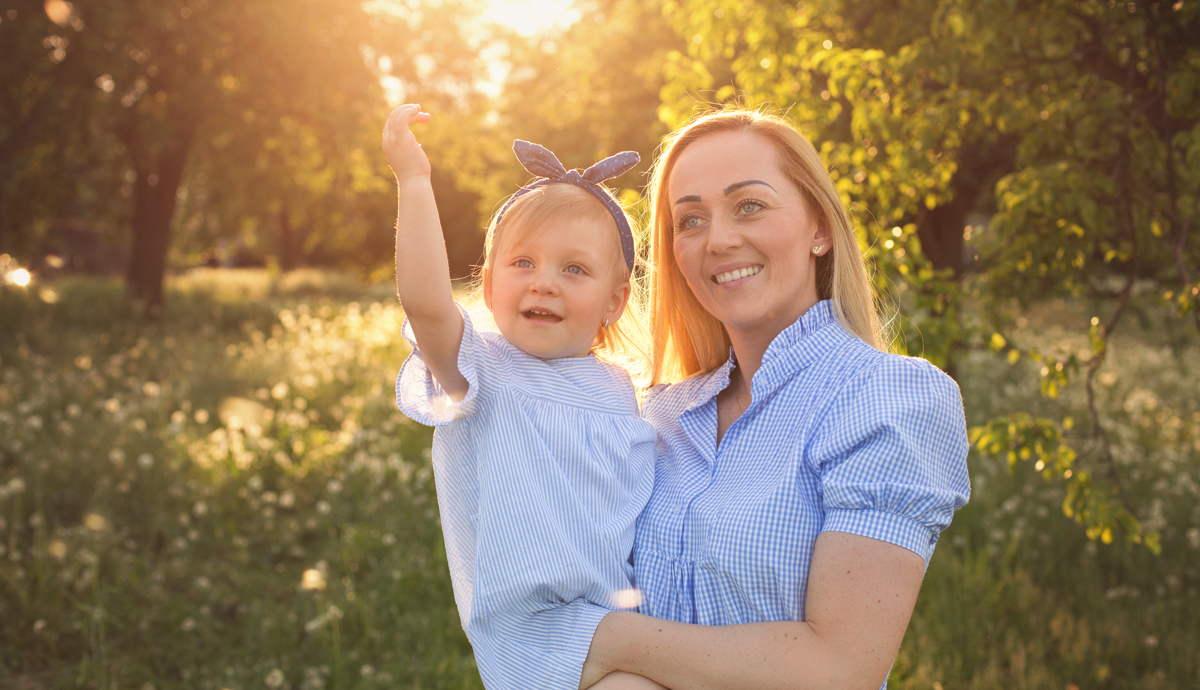 Katarzyna Pawlak - Fotografia Rodzinna - uśmiechnięte mama z córką na rękach na tle zielonego sadu
